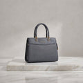 A structured gray leather handbag with double top handles and gold-tone hardware, displayed on a white marble slab against a neutral gray studio background.