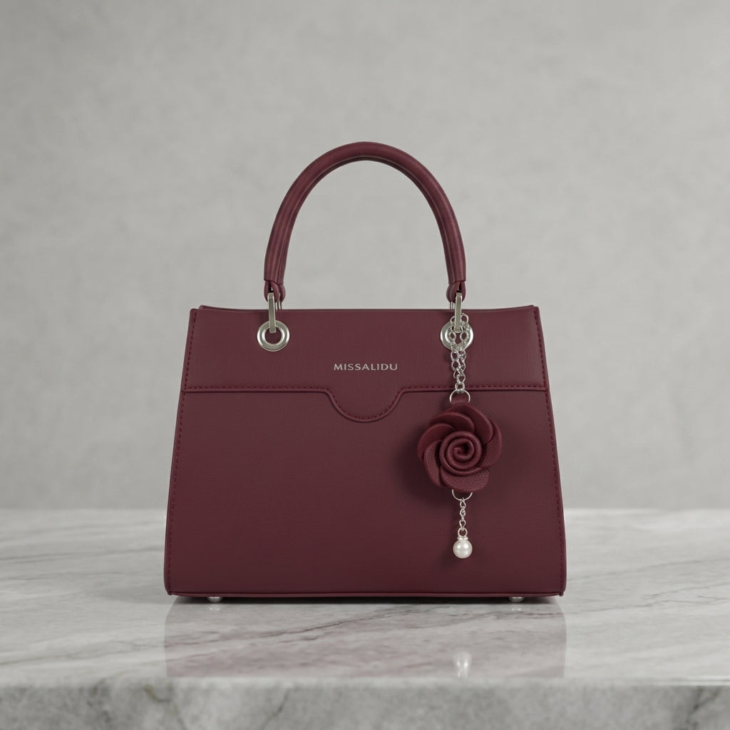 A burgundy leather handbag with silver-tone hardware, featuring a top handle, a decorative rose charm, and the MISSALIDI brand name on the front, displayed on a light gray marble surface against a textured gray background.