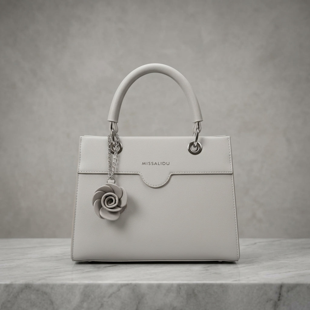 A structured light gray leather handbag with a top handle, silver-tone hardware, and a hanging gray fabric rose charm. The brand name 'MISSALIDU' is debossed on the front flap.