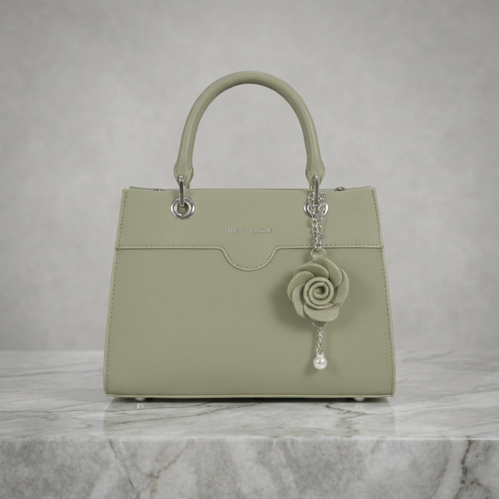 A structured top-handle handbag in a muted sage green color, featuring a matching fabric rose charm with a silver-tone chain and pearl drop, displayed on a light gray marble surface against a textured gray background.
