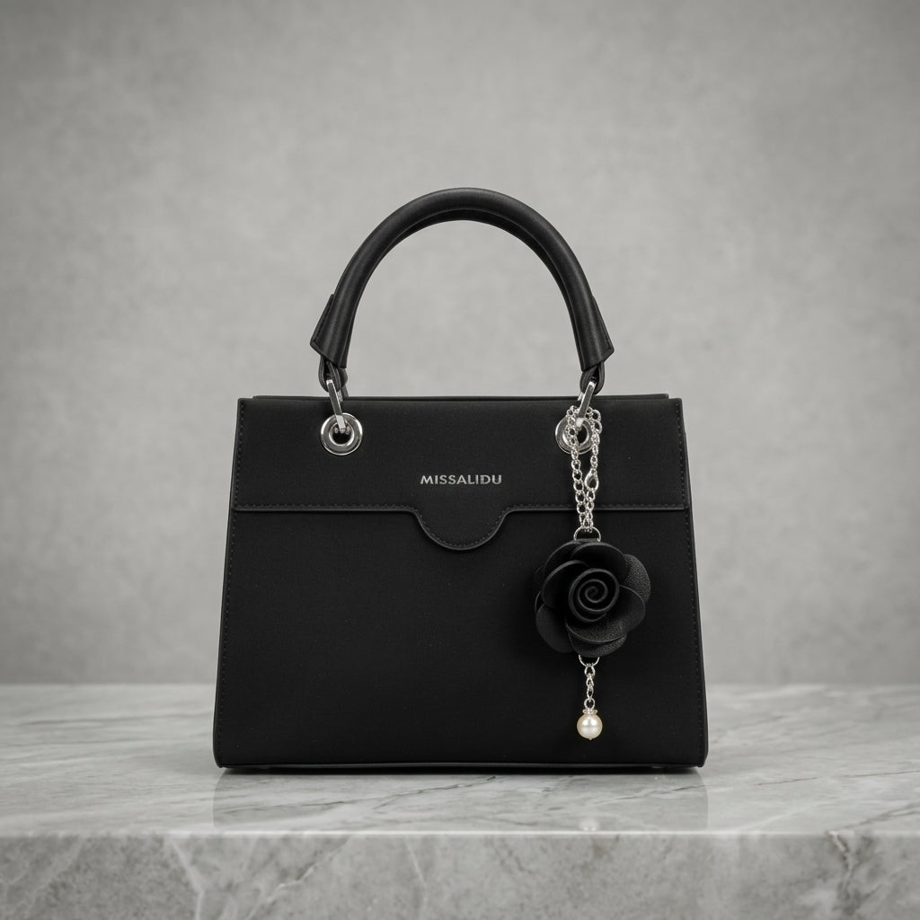 A black MISSALIDU top-handle handbag with silver-tone hardware, featuring a hanging black rose charm with a pearl bead, displayed on a gray marble surface against a textured gray background.