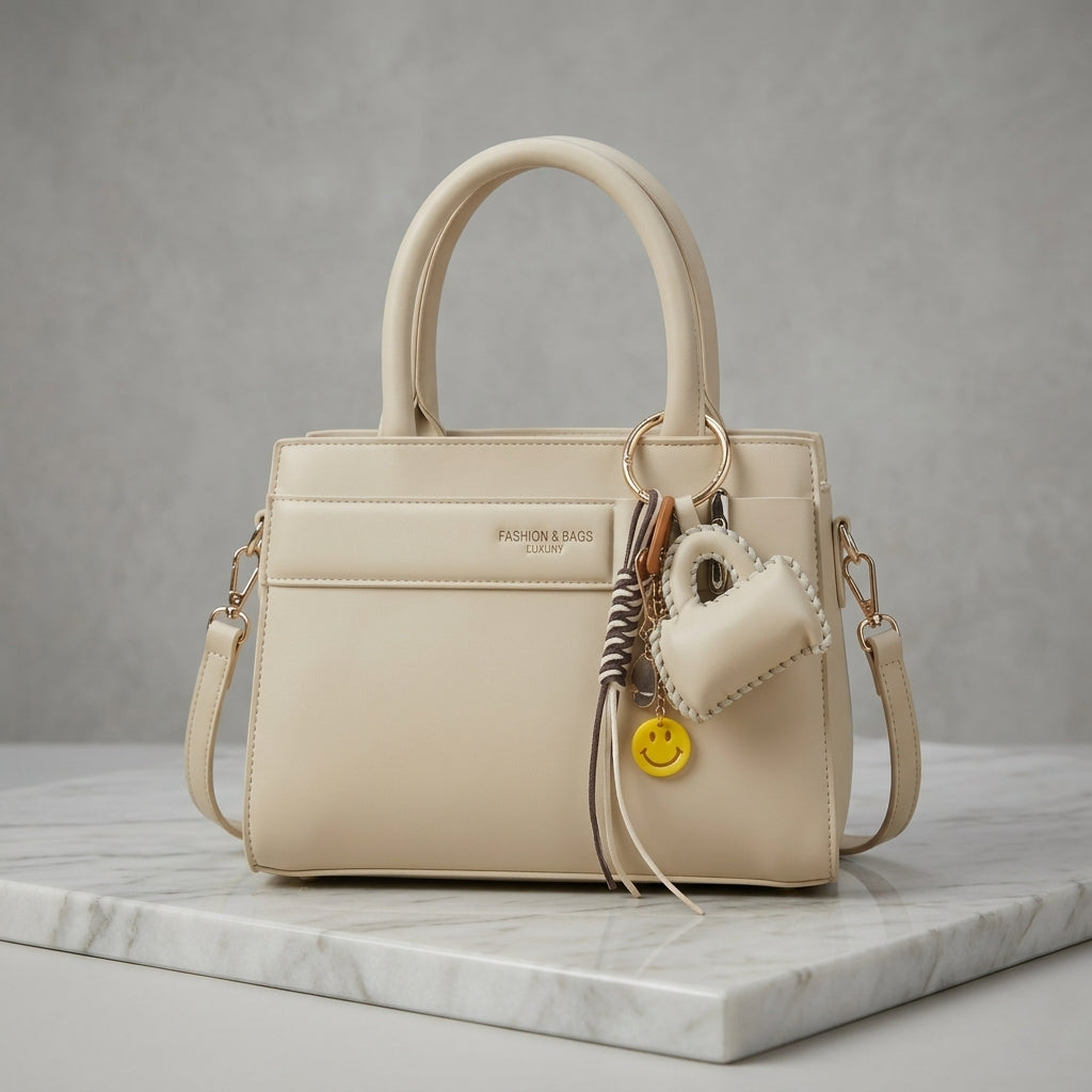A structured cream-colored leather handbag with double top handles, a front pocket, and a hanging charm set including a small leather heart, a yellow smiley face tag, and a striped rope detail, displayed on a white marble slab against a gray background.