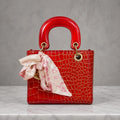 A red crocodile-embossed leather handbag with a glossy top handle and a pink patterned silk scarf tied to the front, displayed on a white marble surface against a textured gray background.