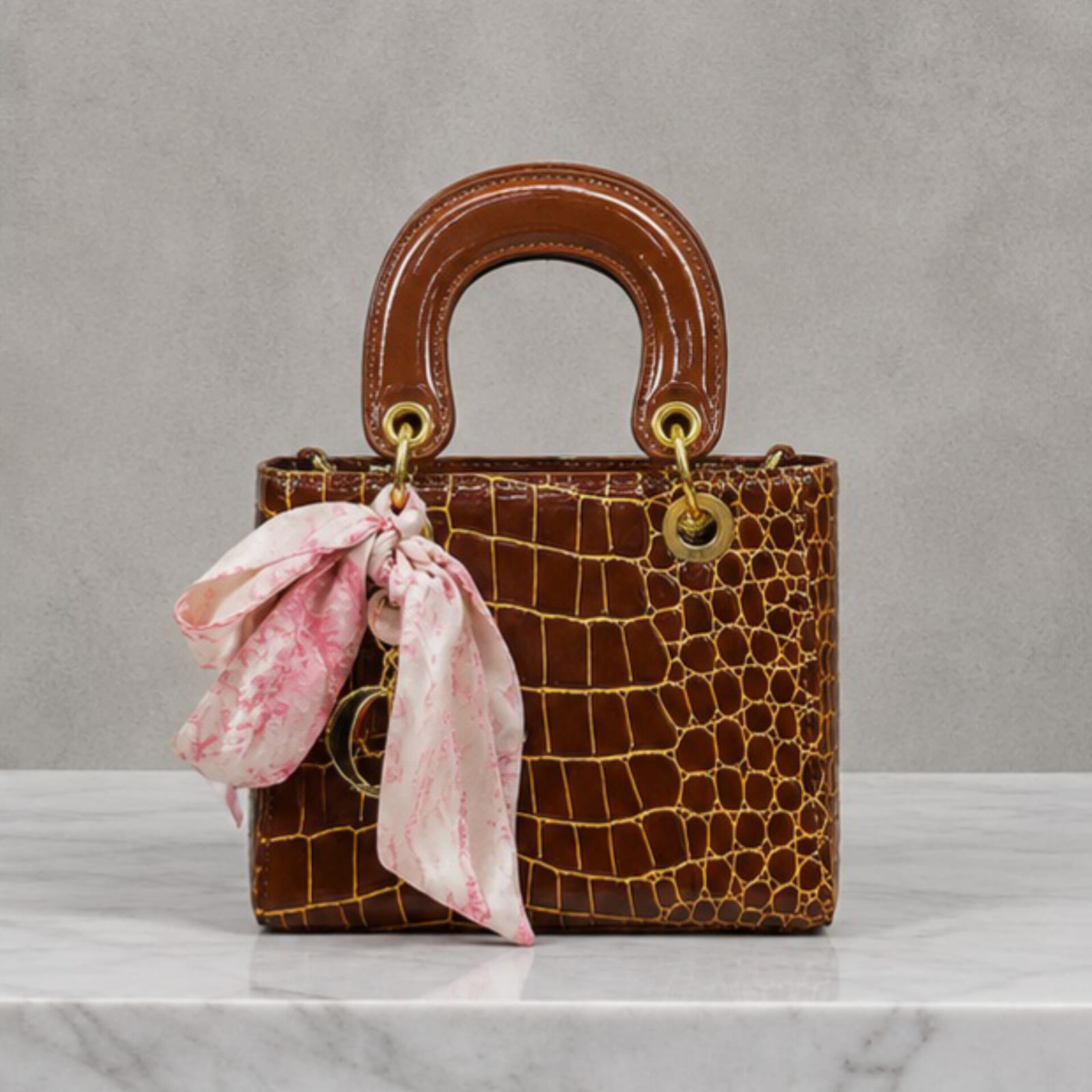 A structured brown crocodile-embossed handbag with a glossy brown top handle, gold-tone hardware, and a pink and white patterned silk scarf tied as a charm, displayed on a white marble surface against a textured gray background.