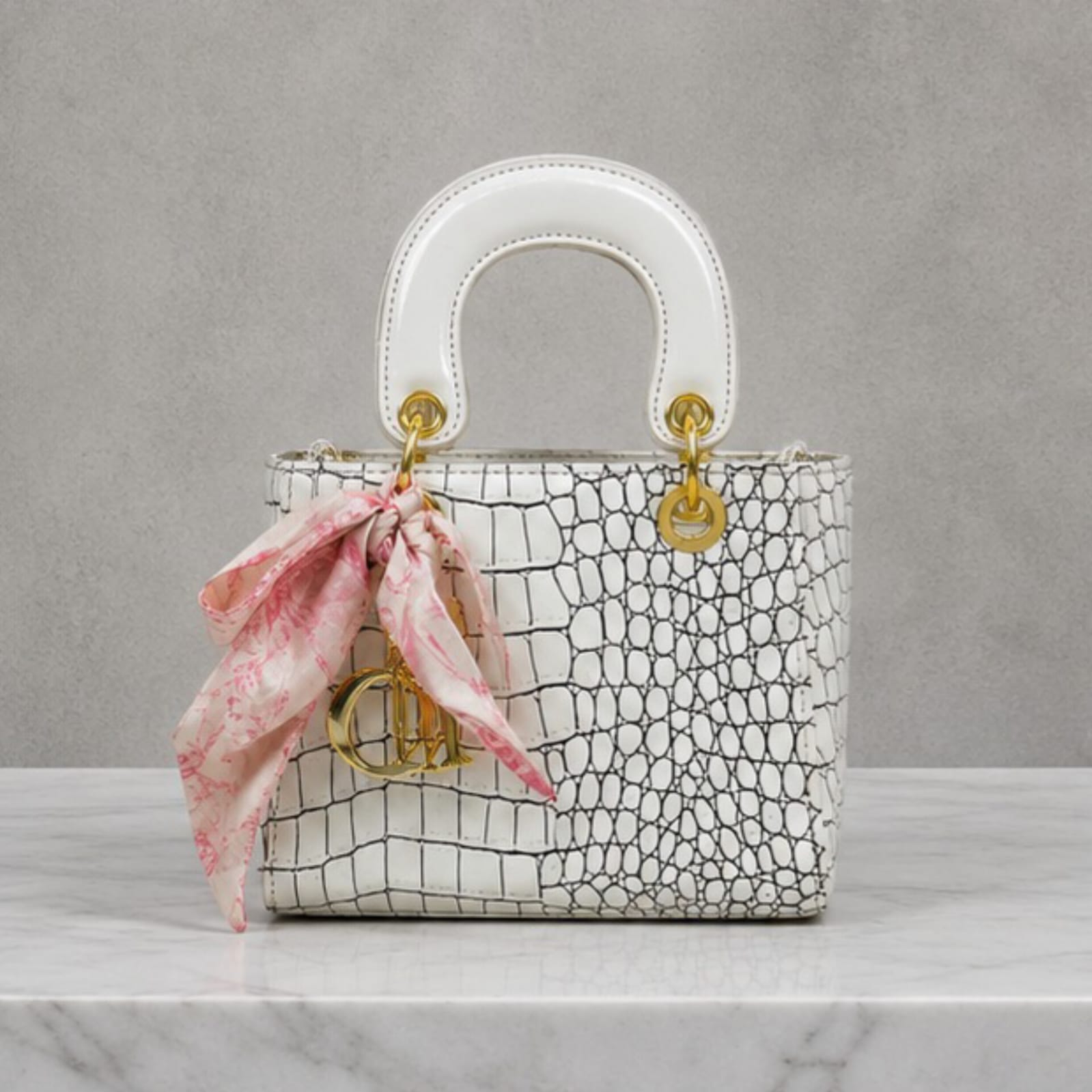 A structured handbag with a white and black crocodile-embossed pattern, featuring a white top handle, gold-tone hardware, and a pink patterned silk ribbon tied in a bow on the front.