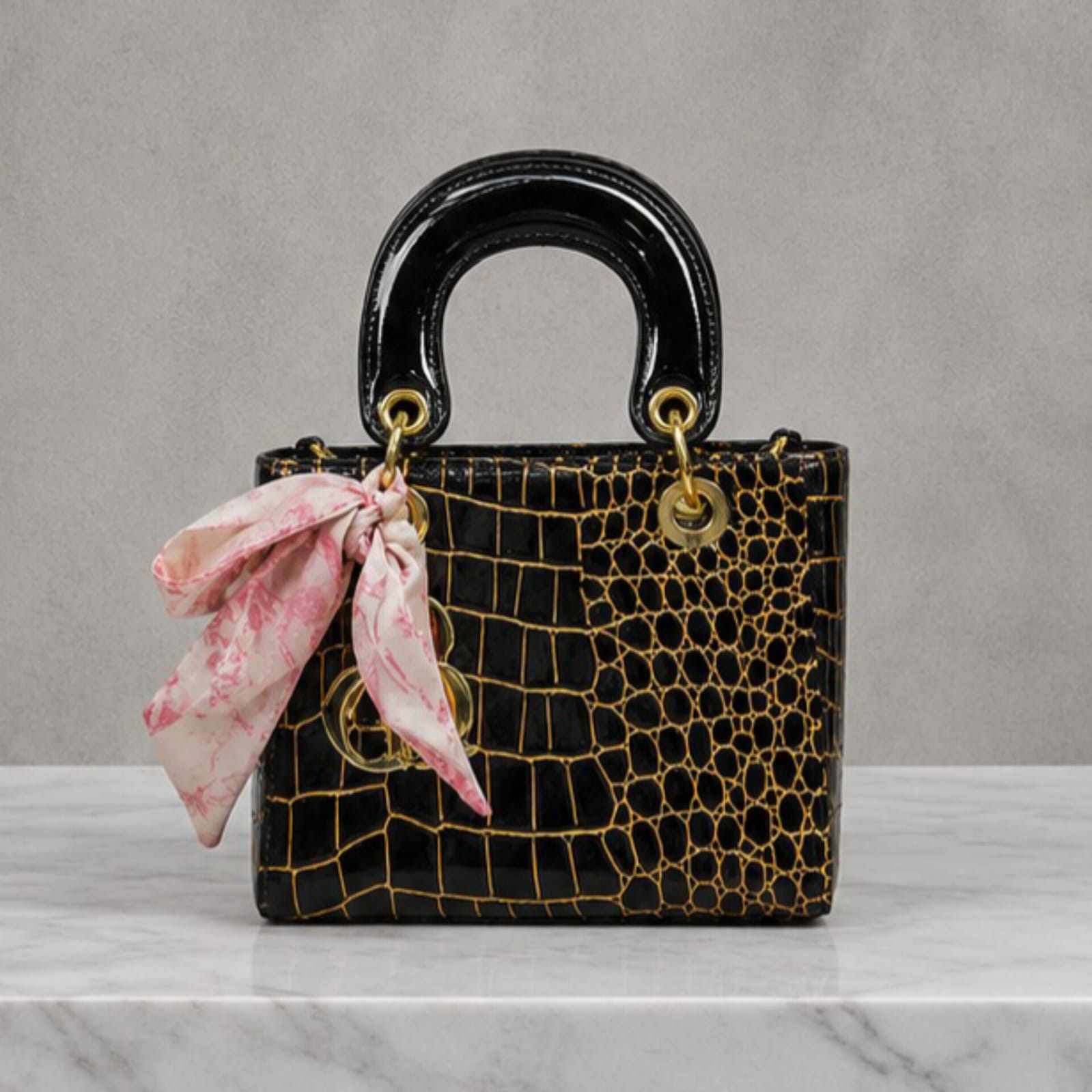 A structured black crocodile-embossed handbag with gold-tone hardware and a pink patterned silk scarf tied to the side, displayed on a white marble surface against a gray background.